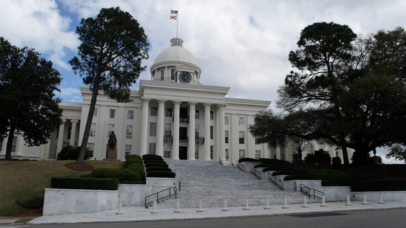 A large limestone building with a clock tower and ivory stairs surrounded by trees on a cloudy day