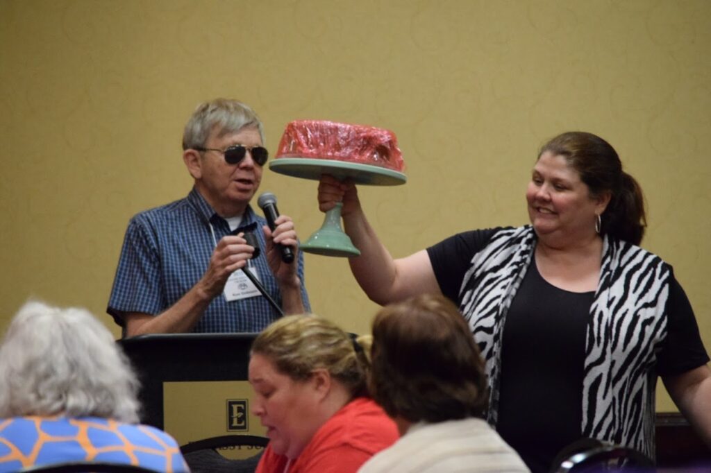 A man with sunglasses uses a mic while a woman carries a pink cake on a green stand