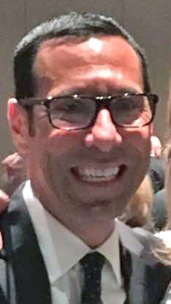 A man with short, dark hair and glasses. He is smiling broadly, showing his teeth. He is wearing a dark suit, a white shirt, and a dark tie. The background is somewhat blurred, but it appears to be an indoor setting with other people present. The lighting is soft and warm.