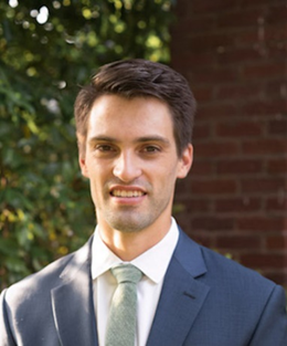 A young man with short, dark brown hair and a light complexion. He is wearing a dark blue suit, a white dress shirt, and a light green tie. He is smiling slightly and standing in front of a background that includes some greenery and a brick wall. The lighting suggests it is taken outdoors, possibly in a garden or park setting.