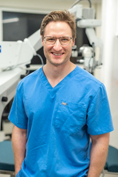 A man standing and smiling at the camera. He is wearing blue medical scrubs and glasses. The background appears to be a medical or clinical setting, with equipment that looks like it could be used for eye examinations or surgeries. The man has short, light brown hair and a friendly expression.