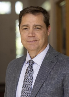 A middle-aged man with short brown hair and a light complexion. He is wearing a gray suit jacket, a light blue dress shirt, and a patterned tie. He has a slight smile on his face and is standing indoors, with a blurred background that includes a doorway and some natural light coming through windows.