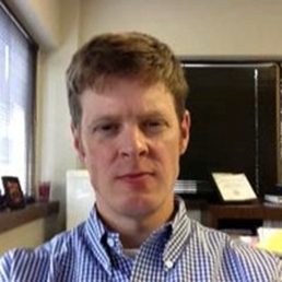 A man with short, light brown hair and fair skin. He is wearing a blue and white checkered button-up shirt. The background appears to be an office setting with a window on the left side, letting in some natural light. There are some office items visible, such as a desk, a computer monitor, and some papers. The man has a neutral expression on his face.