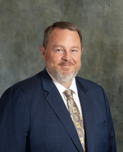 A middle-aged man with short, light brown hair and a neatly trimmed beard. He is wearing a dark blue suit, a white dress shirt, and a patterned tie with shades of gold and blue. The background is a textured, muted greenish-gray color. The man is smiling slightly and looking directly at the camera.