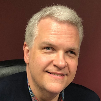 A middle-aged man with short, gray hair. He has a light complexion and is smiling gently. He is wearing a dark blazer over a plaid shirt. The background is a solid, dark maroon color. The man appears to be sitting in an office chair.